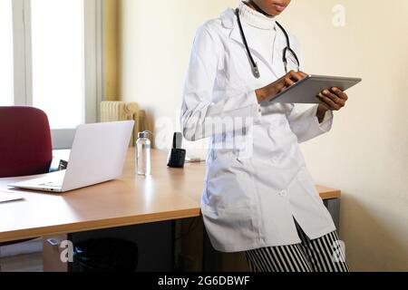 Crop young black female doctor in medical coat with stethoscope working with tablet in modern clinic office Stock Photo