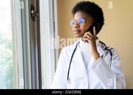 Young African American female doctor in white coat with stethoscope talking mobile phone while standing near window in clinic Stock Photo