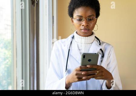 Serious young African American female doctor in white coat with stethoscope checking information on mobile phone while standing near window in clinic Stock Photo
