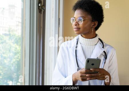 Young African American female doctor in white coat with stethoscope checking information on mobile phone while standing near window in clinic Stock Photo