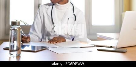 Crop unrecognizable black female physician with stethoscope writing information on paper sheet while preparing medical report at table in office of mo Stock Photo