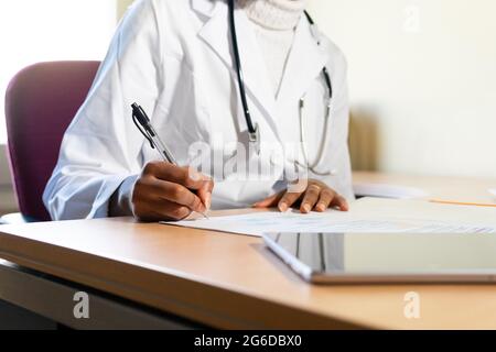 Crop unrecognizable black female physician with stethoscope writing information on paper sheet while preparing medical report at table in office of mo Stock Photo