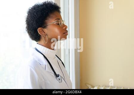 Side view of competent young African American female physician in white medical coat and eyeglasses with stethoscope closed eyes while standing in cli Stock Photo