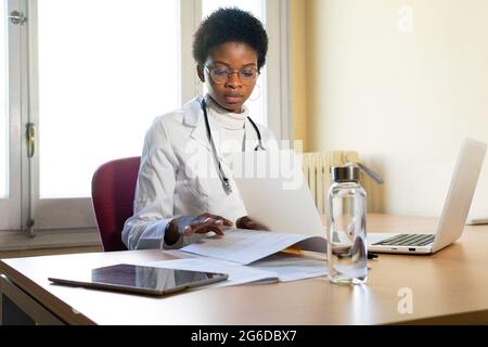 Young African American female physician in white robe with stethoscope sitting at table with laptop and reading medical records while working in clini Stock Photo