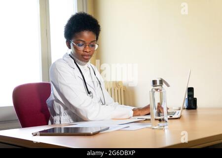 Young African American female physician in white robe with stethoscope sitting at table with laptop and reading medical records while working in clini Stock Photo
