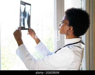 Side view of young black female medic standing near window and examining x ray scan while working in clinic Stock Photo