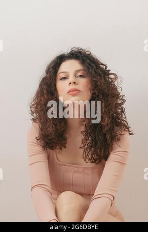 Gentle dreamy female with curly hair and in bodysuit sitting on floor against white wall in studio Stock Photo