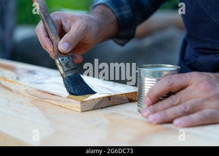 Crop anonymous male carpenter with paintbrush applying lacquer on wooden board while working in professional woodworking workshop Stock Photo