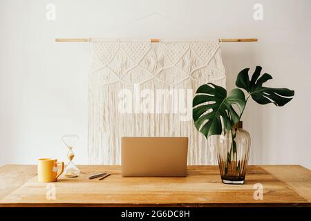 Laptop and smartphone on wooden table with cup of tea and glass vase with green plants against white wall Stock Photo