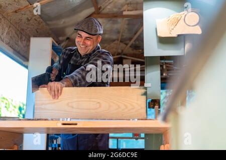 Happy carpenter using sharp saw while cutting lumber plank at workbench in professional workshop Stock Photo