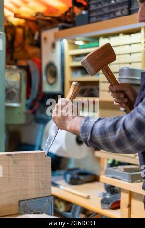 Side view of crop male master using hammer and chisel while creating wooden detail in carpentry workshop Stock Photo