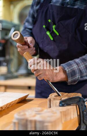 Unrecognizable male woodworker with wooden hammer and chisel carving wooden detail while working in professional joinery workshop Stock Photo