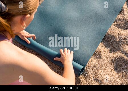 From above cropped unrecognizable young female in sportswear placing yoga mat on sand while preparing for practice on beach near ocean Stock Photo