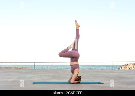 Side view of fit female doing yoga in Salamba Sirsasana while balancing on mat near sea Stock Photo