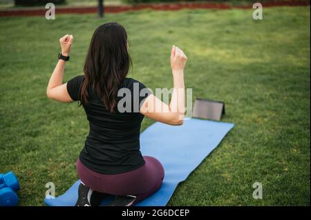 High angle back view of female athlete warming up body and watching online video on tablet while training on lawn in park Stock Photo