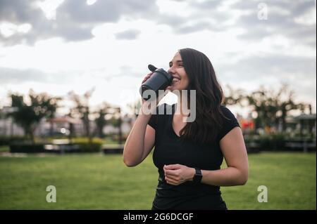 Smiling Latin female athlete drinking refreshing water during workout on meadow in park Stock Photo