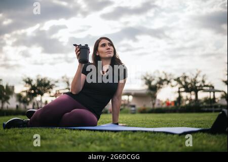 Ethnic athletic female sitting on mat on lawn and taking break during workout while drinking water Stock Photo