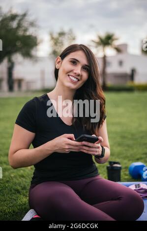 Smiling Latin female athlete browsing mobile phone while sitting on mat during training on meadow and looking at camera Stock Photo