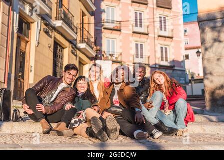 Company of content multiracial friends sitting in street and looking at camera on sunny day Stock Photo