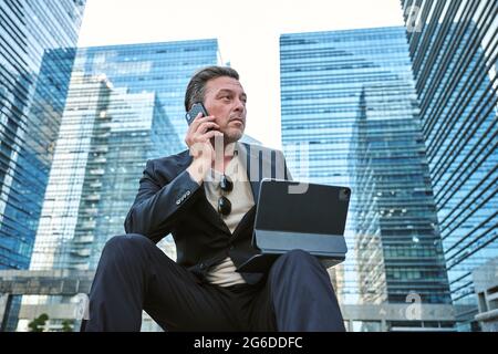 Business man having a call on his smartphone and working with his tablet while sitting around office's buildings Stock Photo