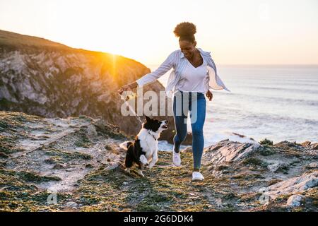 Young African American female owner running with Border Collie dog while spending time together on beach near waving sea at sunset Stock Photo