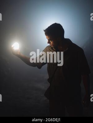 Young male speleologist with bright flashlight in hand exploring dark subterranean cave during extremal adventure Stock Photo