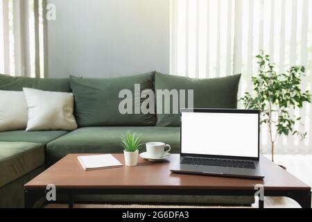 Work from home, workspace, desktop, remote work concept, gray thin laptop computer on brown wooden table with white cup of coffee, green sofa, flowerp Stock Photo