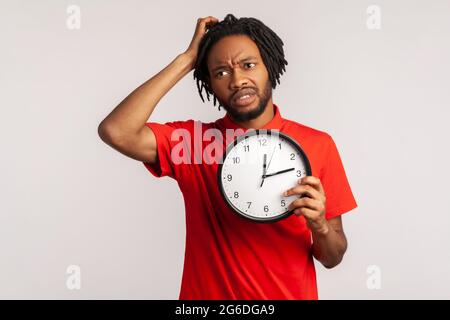 Confused man with dreadlocks wearing red T-shirt, rubbing his head holding in hands big wallclock, has no time , worried about deadline, looking away. Stock Photo