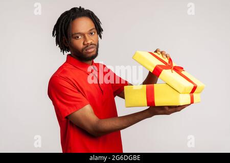Disappointed man with dreadlocks in red casual T-shirt, holding unpacked gift box, has frustrated dissatisfied facial expression, upset with present. Stock Photo