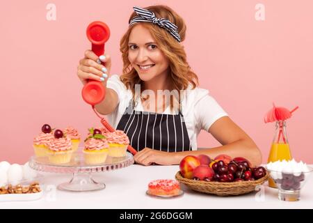 Smiling blonde woman baker in apron sitting at table surrounded with ready-made pastry and fruits, showing handset to camera and smiling. Indoor studi Stock Photo
