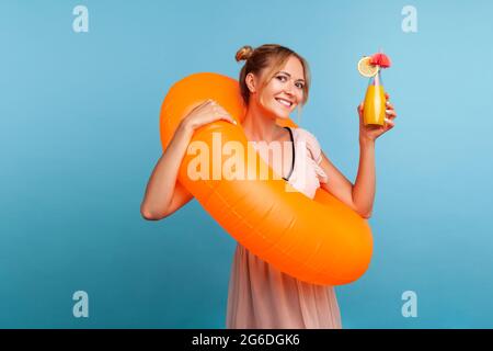 Happy blonde woman with orange rubber ring rising hand with cocktail and looking directly at camera with charming smile, wearing summer dress. Indoor Stock Photo