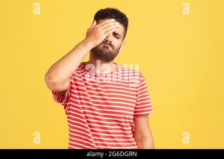 Unhappy forgetful man with beard in striped t-shirt making facepalm gesture keeping hand on head, blaming himself for bad memory, unforgivable mistake Stock Photo