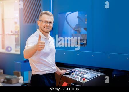 Engineer young man in glasses operates CNC automatic metalworking lathe or laser, industrial worker in modern factory. Stock Photo