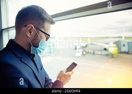 Traveler young man business with face mask waiting to board on airplane, use phone for online check plane in airport. Concept passenger traveling by c Stock Photo