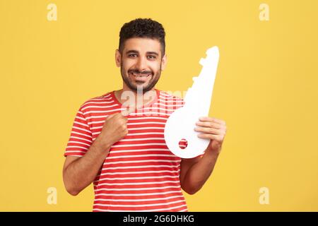 Smiling positive bearded man in striped t-shirt pointing finger at big paper key in his hand, satisfied with renting or purchase of housing. Indoor st Stock Photo