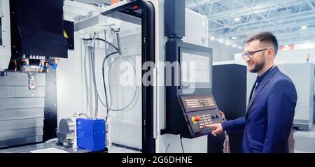 Engineer young man in glasses operates CNC automatic metalworking lathe or laser, industrial worker in modern factory. Stock Photo