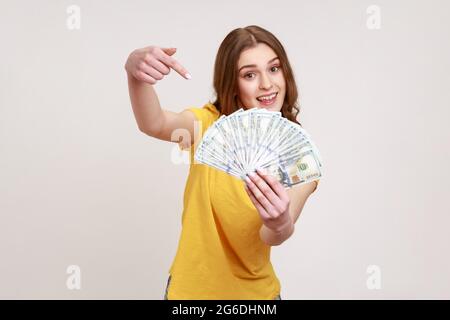 Portrait of joyful rich girl in yellow casual T-shirt holding lot of money, pointing at dollar bills in hand and smiling excitedly, enjoying lottery w Stock Photo