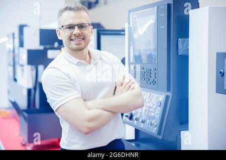Engineer young man in glasses operates CNC automatic metalworking lathe or laser, industrial worker in modern factory. Stock Photo