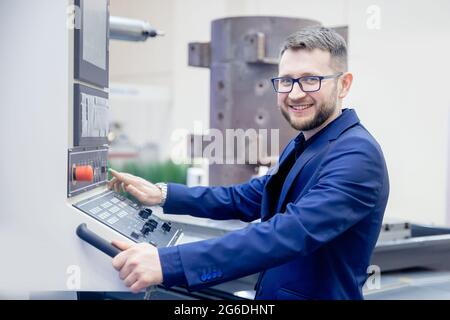 Engineer young man in glasses operates CNC automatic metalworking lathe or laser, industrial worker in modern factory. Stock Photo