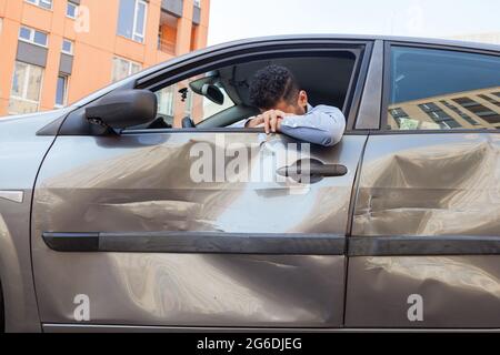 Despaired man sitting behind the wheel of crashed car and puts his head down, crying, broke his car due to traffic violence, scratches and dents on au Stock Photo