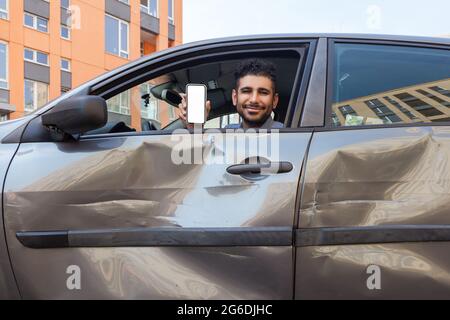 Positive smiling bearded man sitting in dented car and showing mobile phone with empty screen to camera, offers insurance inspection services after ro Stock Photo