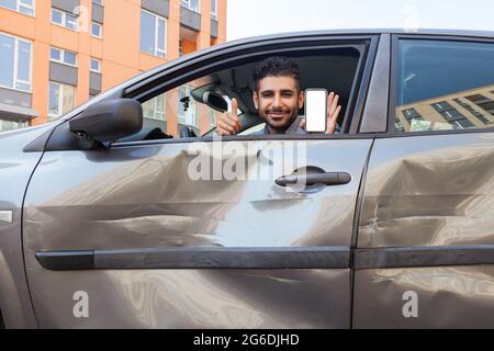 Portrait of bearded man in crashed car looking at camera and smiling, showing thumb up and smart phone with blank screen, advertising insurance inspec Stock Photo