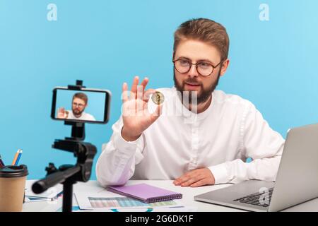 Bearded man blogger holding in hands and showing on smartphone camera golden bitcoin, explaining how to invest in cryptocurrency, mining. Indoor studi Stock Photo