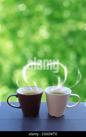 Two cups of green tea with heart shaped steam on a wooden table against green foliage bokeh Stock Photo