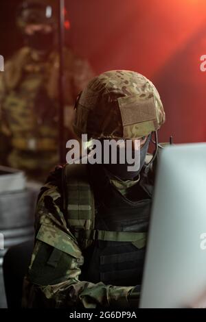 Man in mask, helmet and camouflage uniform committing cyber crime in front of computer Stock Photo