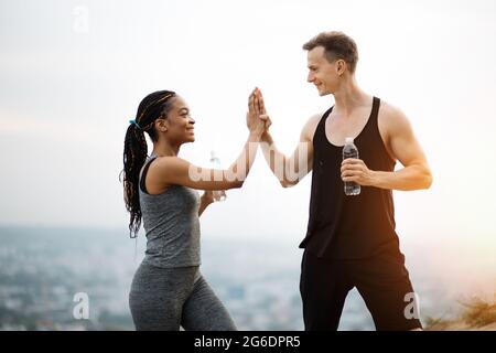 Smiling young couple dressed in sport clothes giving high five to each other after active workout on fresh air. African woman and caucasian man holding bottle of fresh water. Stock Photo