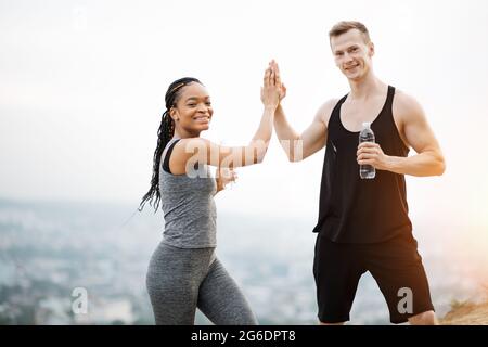 Multicultural couple wearing sport clothes posing with bottle of water in hands outdoors. Caucasian man and black woman giving high five. Active and healthy lifestyles. Stock Photo