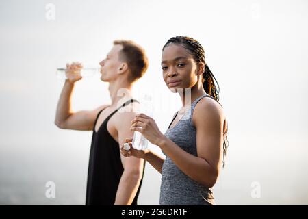 Healthy and fit multicultural couple in sport outfit drinking fresh water from bottle outdoors. Strong man and woman taking break during training on nature. Focus on African woman posing at camera Stock Photo