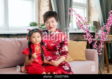 Mother and daughter in red traditional Chinese dresses sitting on couch Stock Photo
