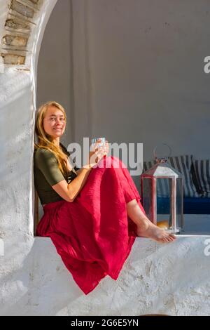 Young woman holding cup and sitting on wall, Greece Stock Photo